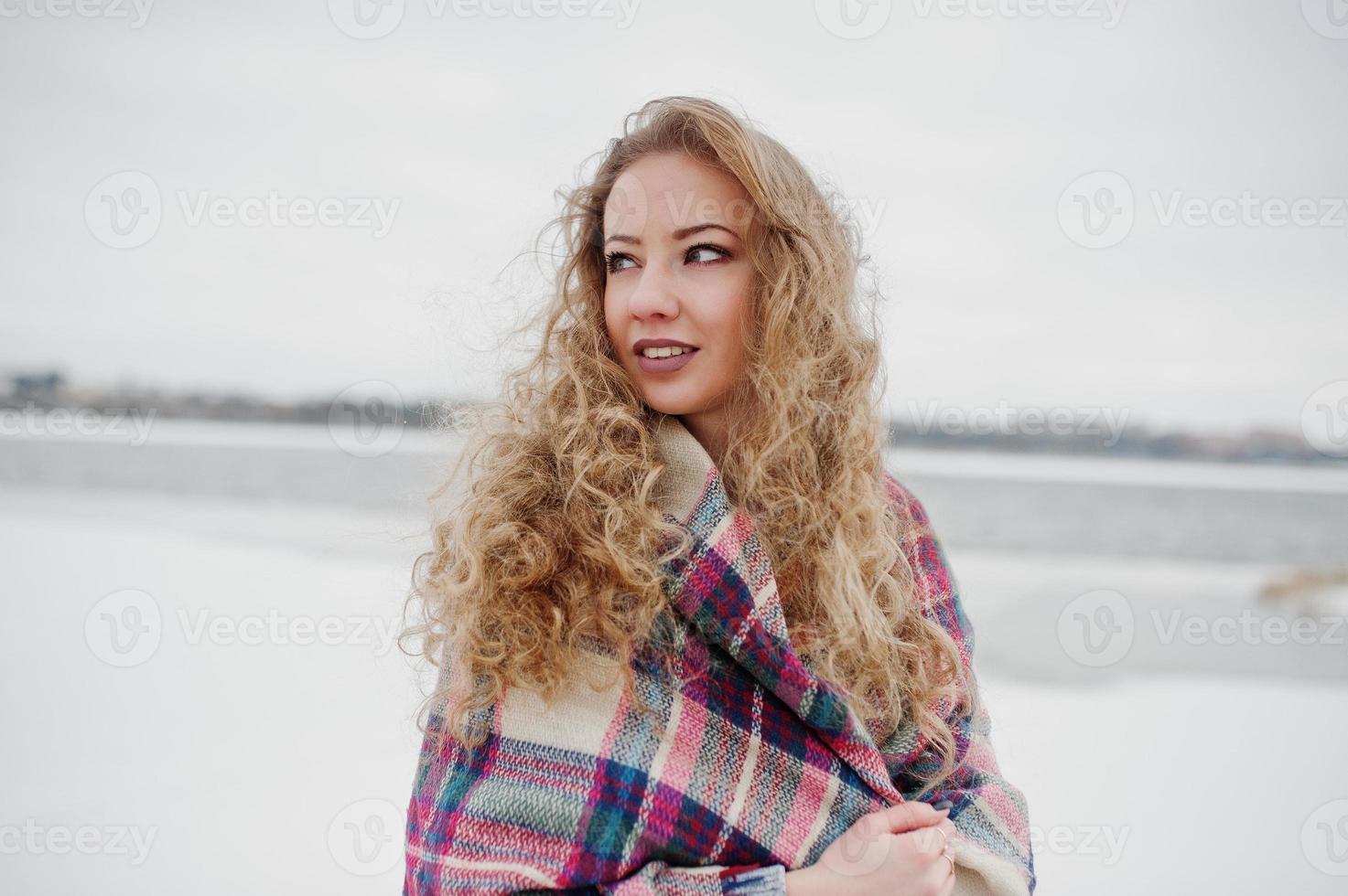 Curly blonde girl in checkered plaid against frozen lake at winter day. photo