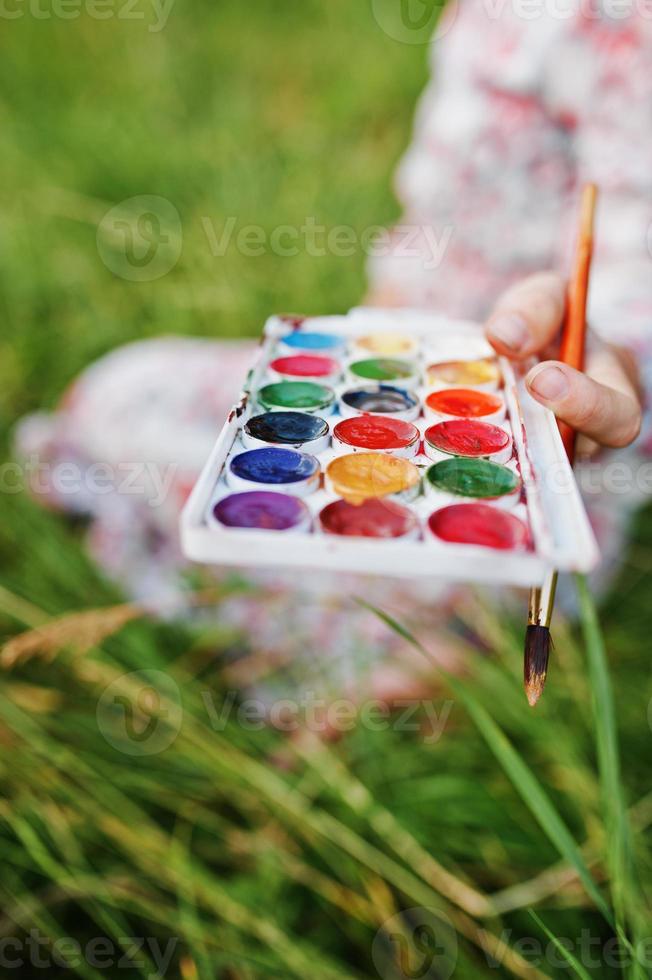 Close-up photo of female hands holding watercolor paints and a brush while painting in nature.