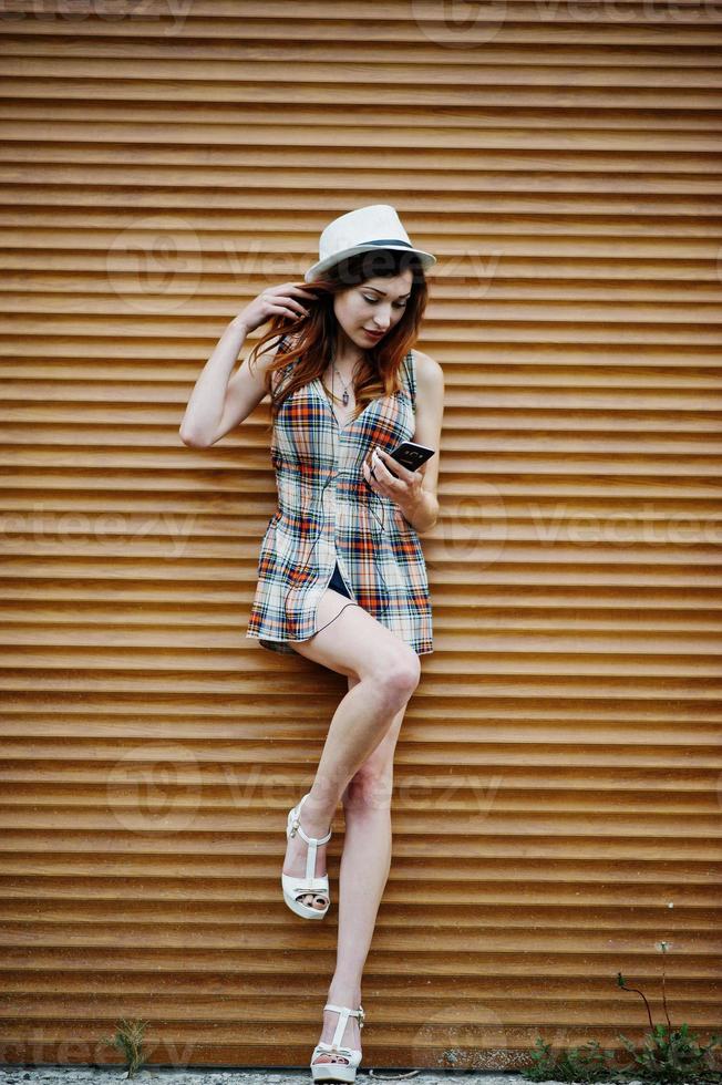 Amazing long legs with hig heels girl wear on hat against shutter listening music from headphones of mobile phone. photo