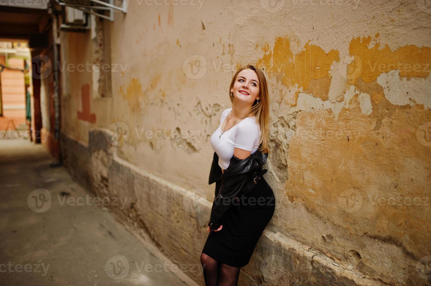 Elegant blonde girl wear on black leather jacket posing at streets of town background old wall with shadows. photo