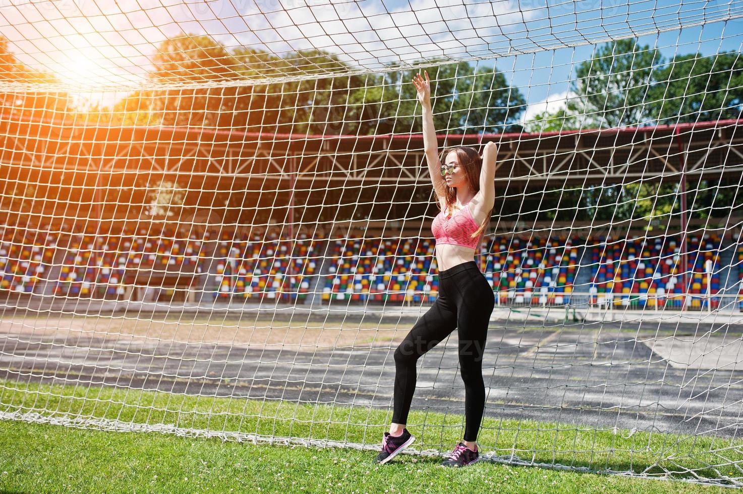 Fitness sporty girl in sportswear and sunglasses posed at stadium football gates outdoor sports. Happy sexy woman. photo