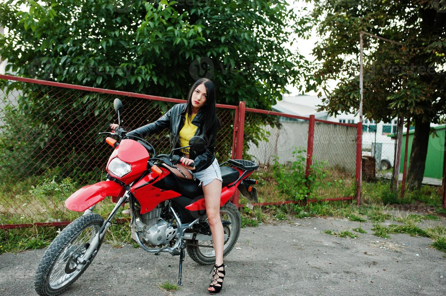 retrato de una mujer genial e impresionante con vestido y chaqueta de cuero negro sentada en una moto roja genial. foto