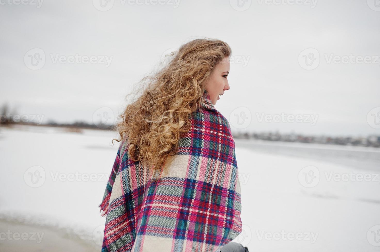 Curly blonde girl in checkered plaid against frozen lake at winter day. photo