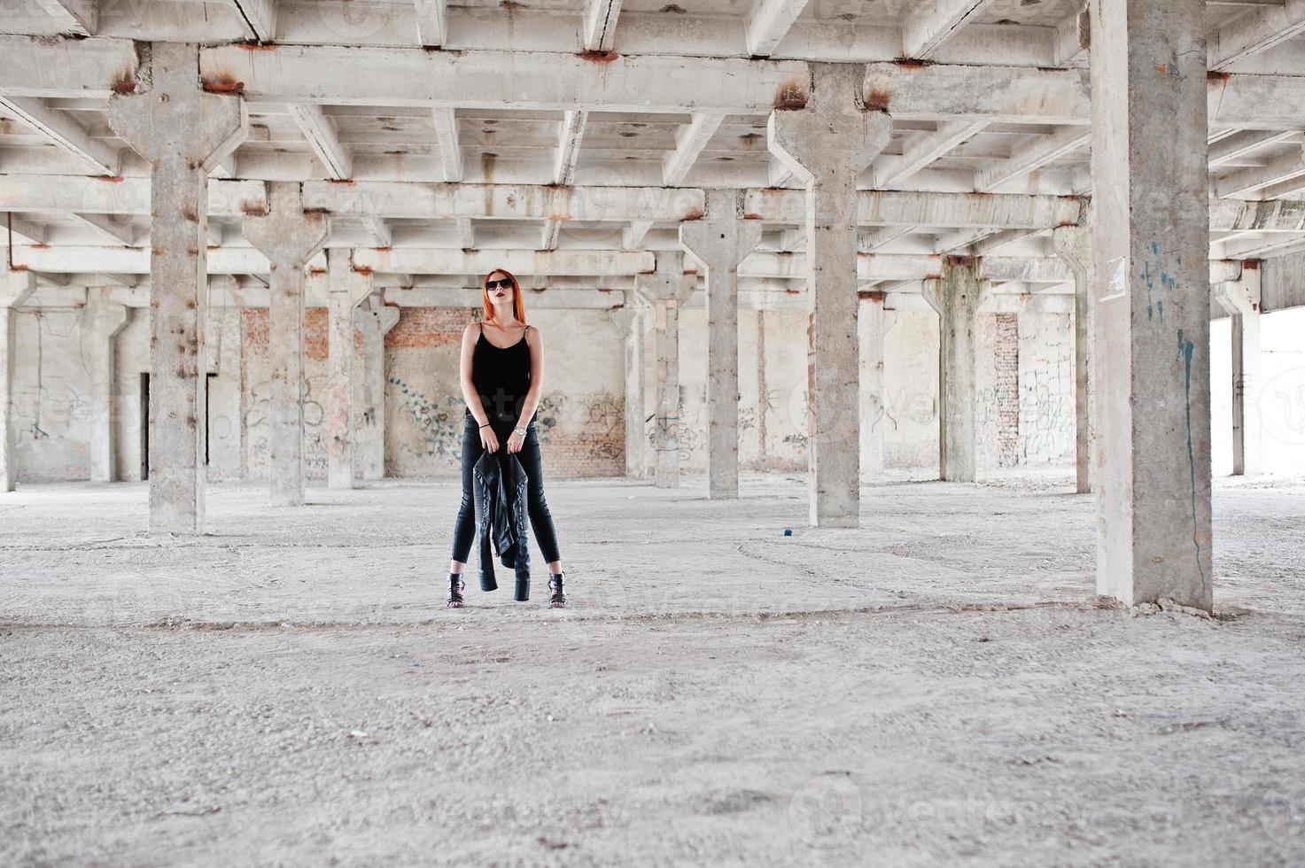 Red haired stylish girl in sunglasses wear in black, against abadoned place. photo