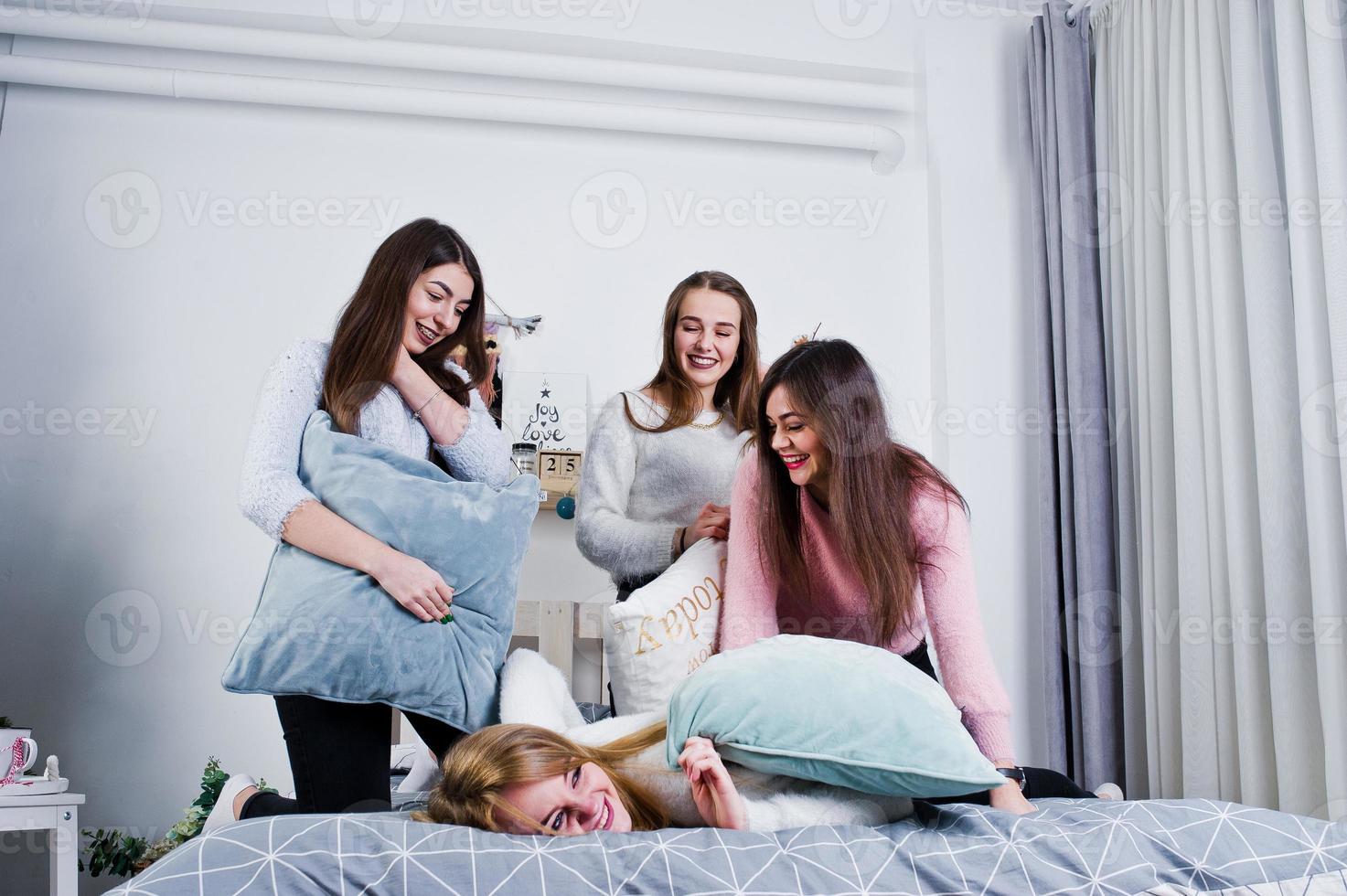 Four cute friends girls wear on warm sweaters and black pants on bed at new year decorated room on studio, play with pillows. photo