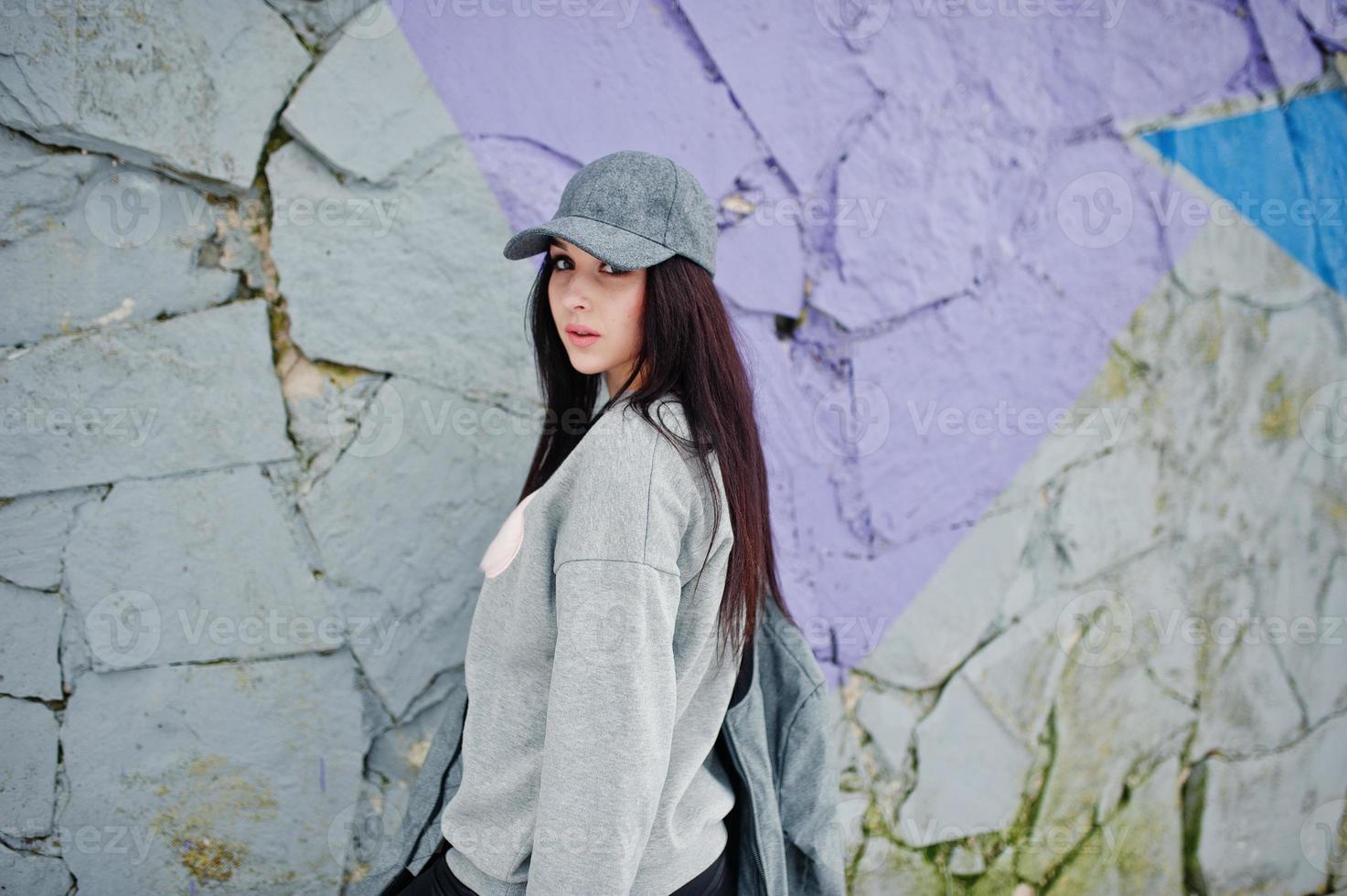 Stylish brunette girl in gray cap, casual street style on winter day against colored wall. photo