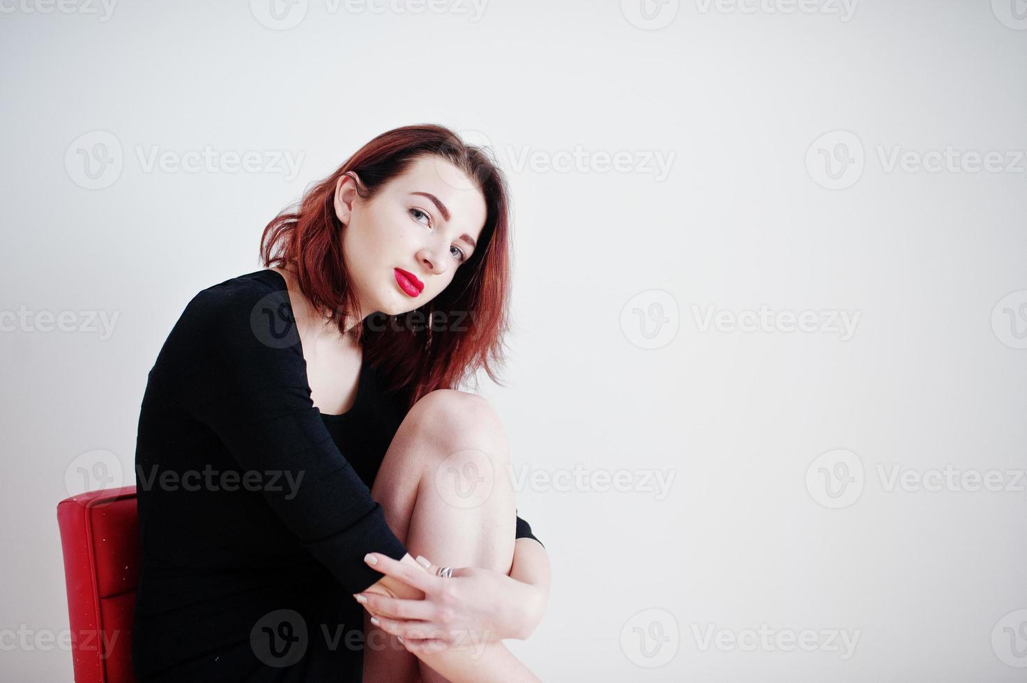 Red haired girl on black dress tunic sitting on red chair against white wall at empty room. photo