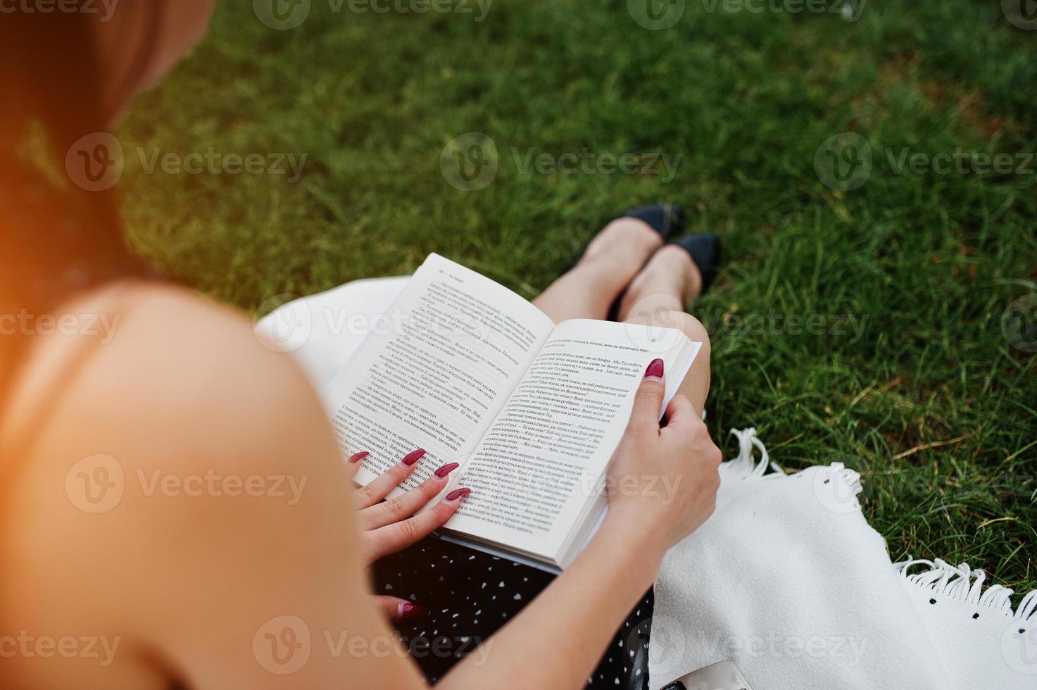 Close-up photo of woman's back while sitting on the blanket on the grass and reading a book.