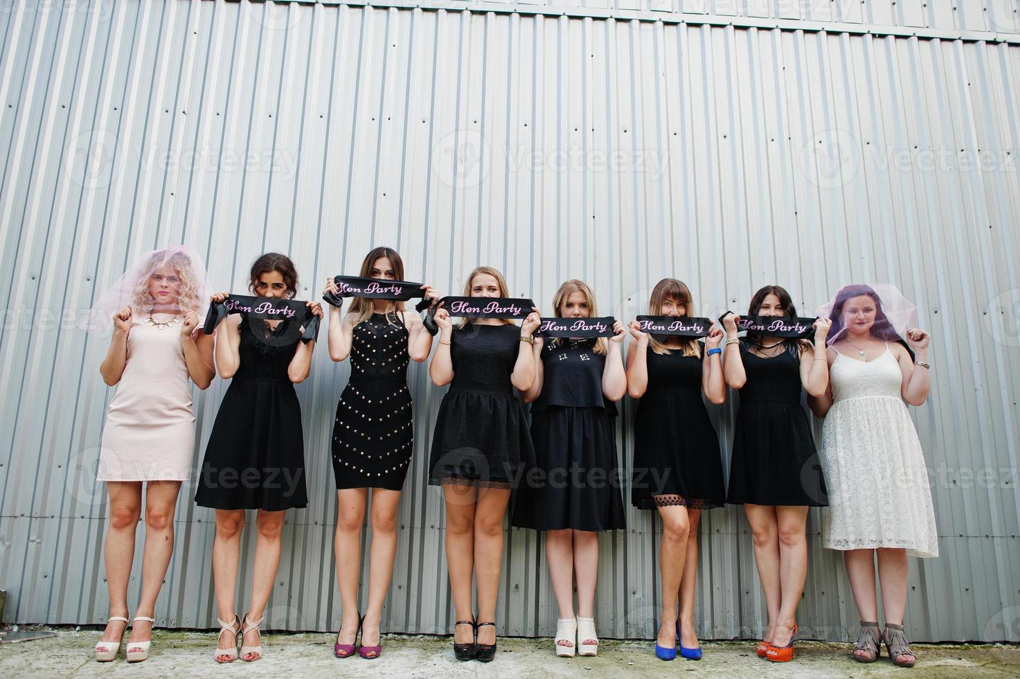 Group of 8 girls wear on black and 2 brides at hen party posed against steel wall. photo