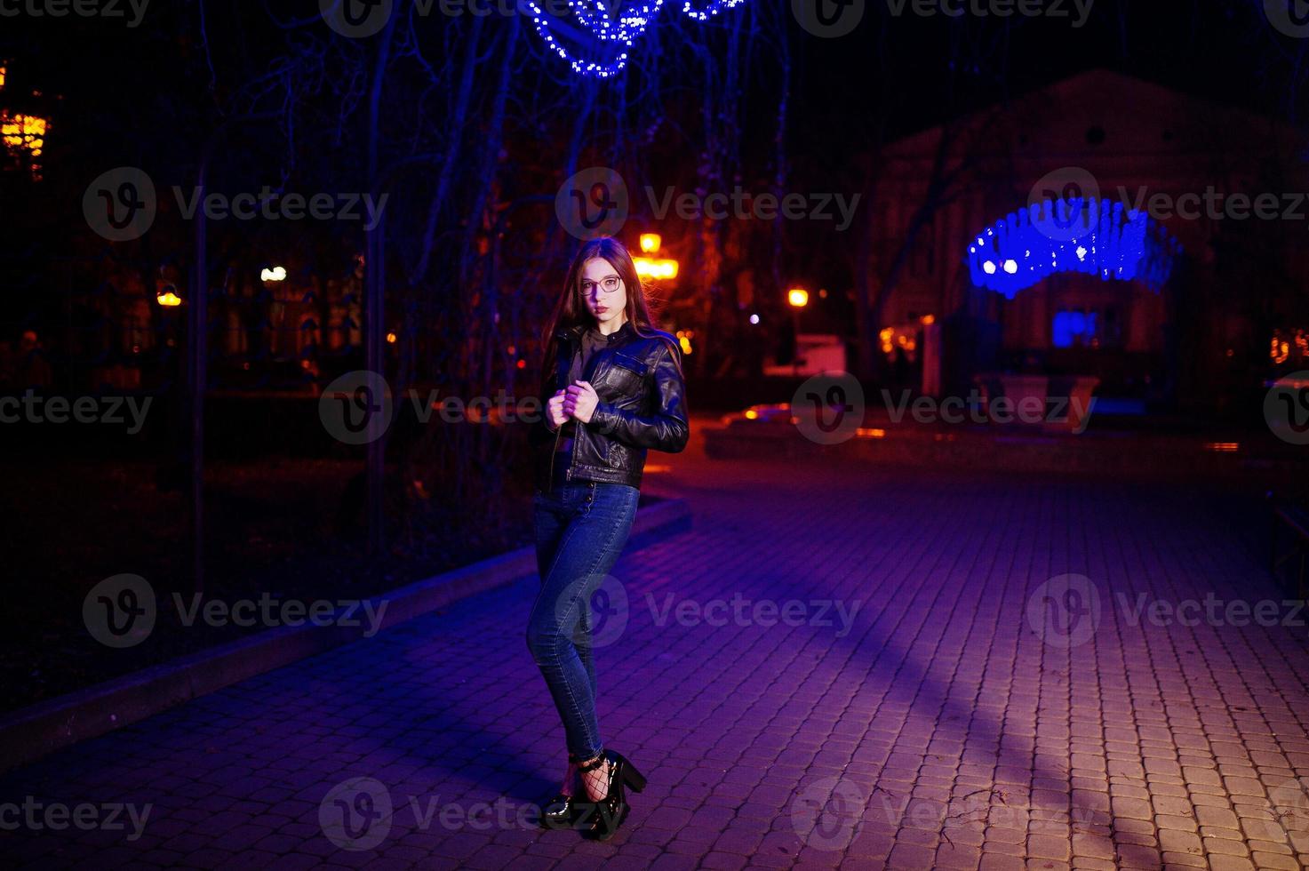 Night portrait of girl model wear on glasses, jeans and leather jacket against blue lights garland of city street. photo