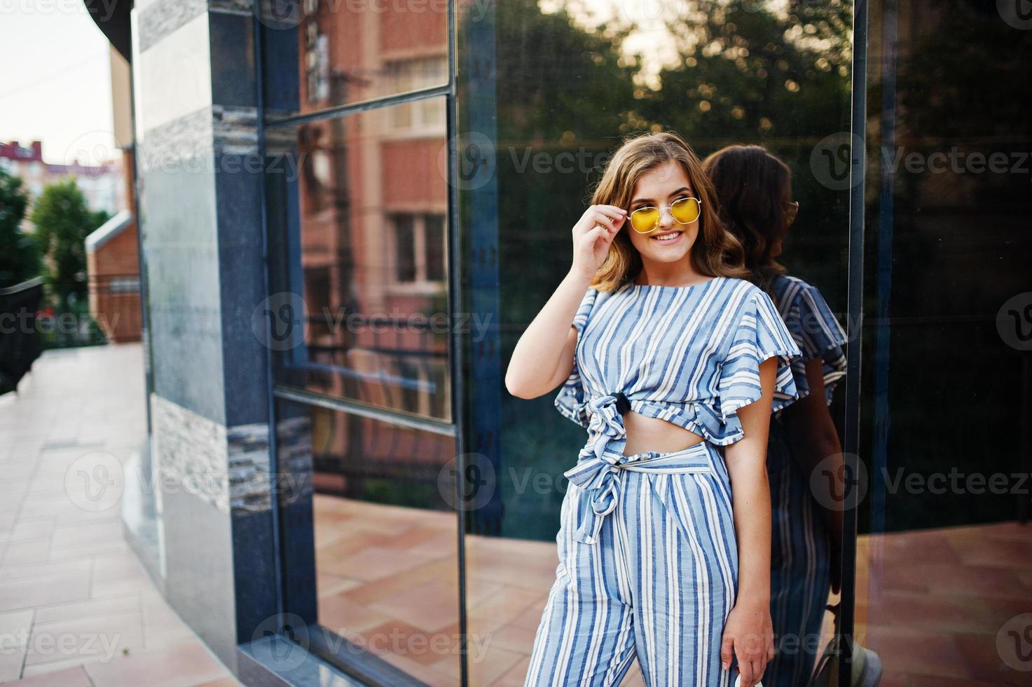 Portrait of a perfect young woman wearing striped overall and yellow sunglasses poses on a balcony of a modern building in a town. photo