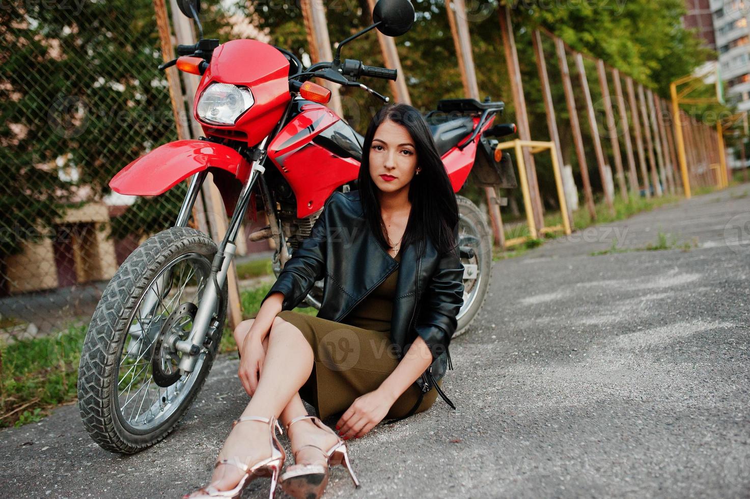 retrato de una mujer genial e impresionante con vestido y chaqueta de cuero negro sentada en una moto roja genial. foto