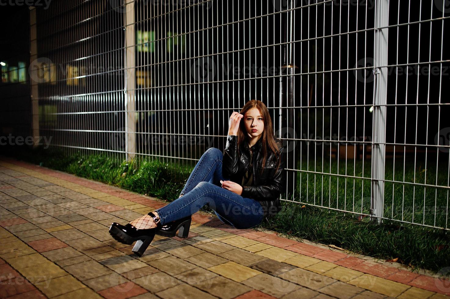 Night portrait of girl model wear on jeans and leather jacket against iron fence. photo