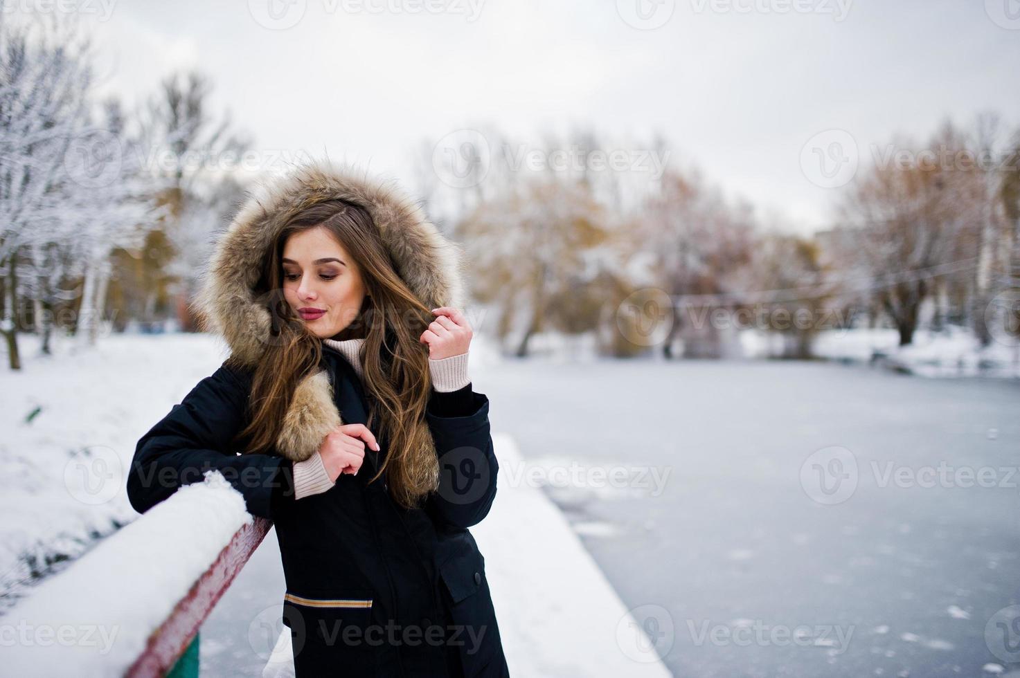 Beautiful brunette girl in winter warm clothing. Model on winter jacket against frozen lake at park. photo