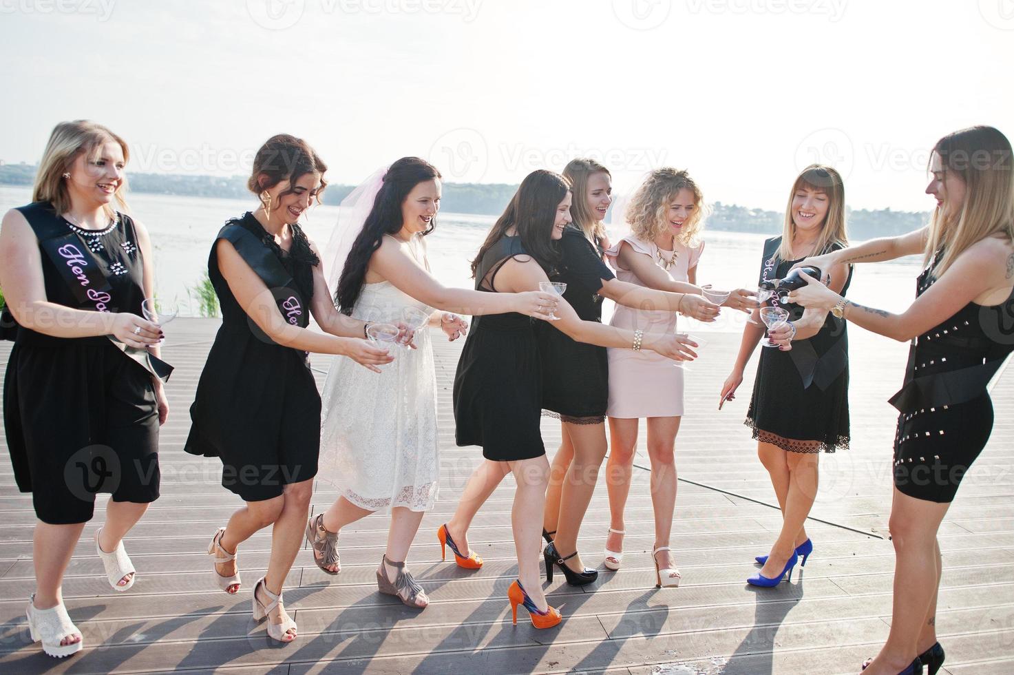 grupo de 8 chicas visten de negro y 2 novias en despedida de soltera contra la playa soleada bebiendo champán. foto