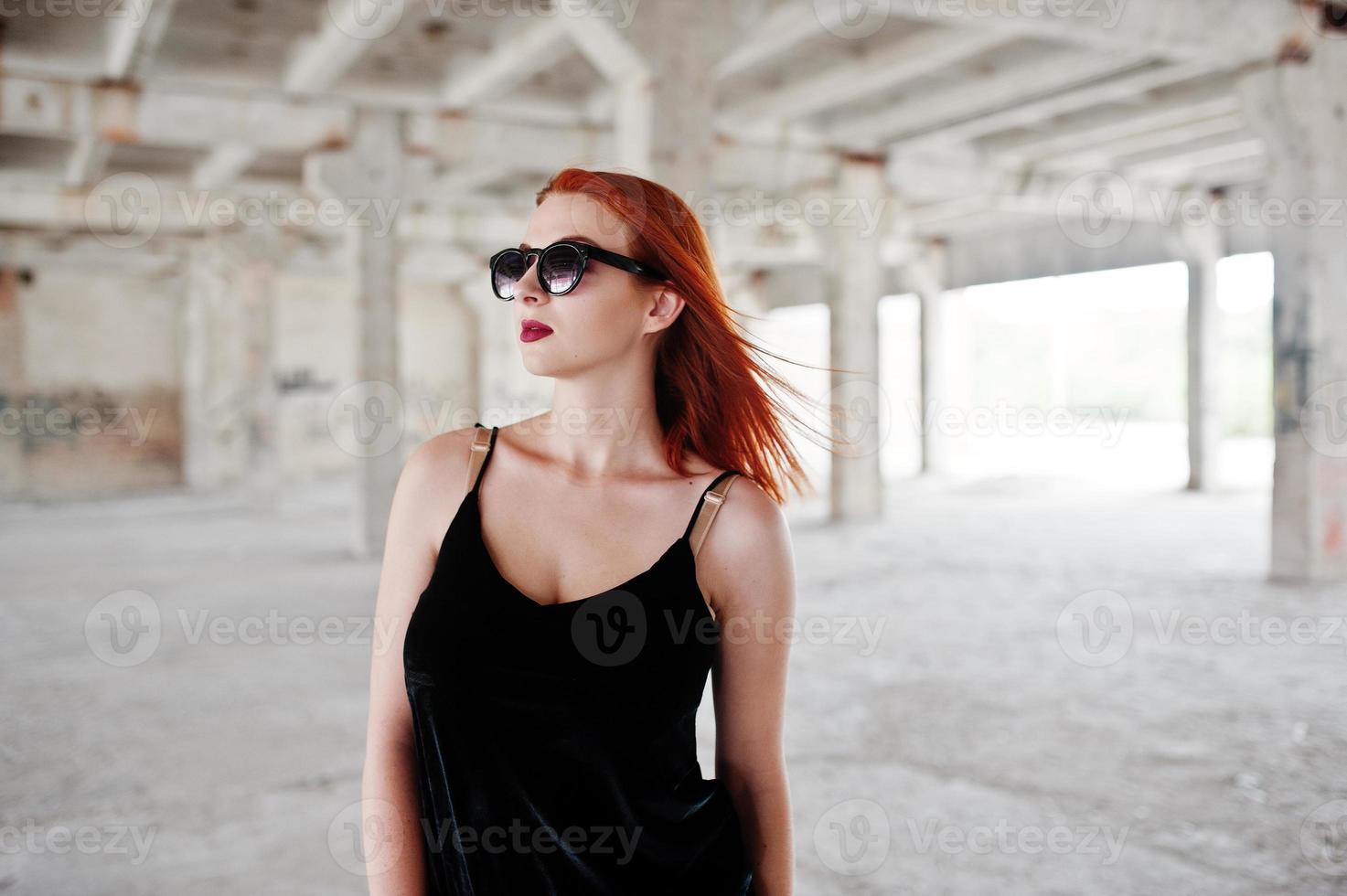 chica elegante de pelo rojo con gafas de sol vestidas de negro, contra un lugar abandonado. foto