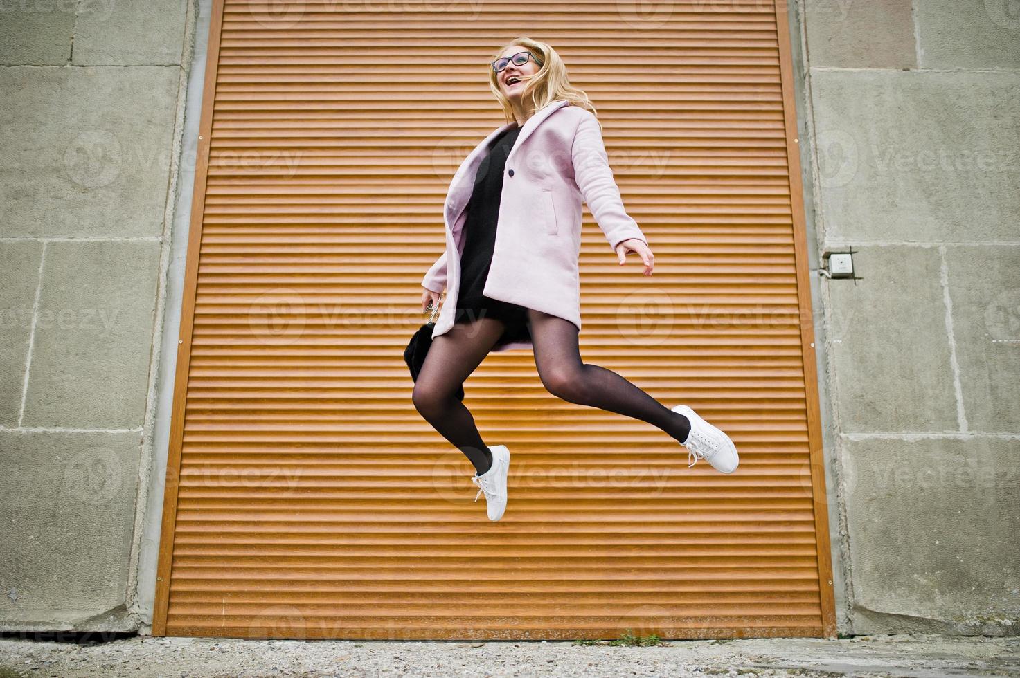 Blonde girl at glasses and pink coat with handbag jump against shutters. photo