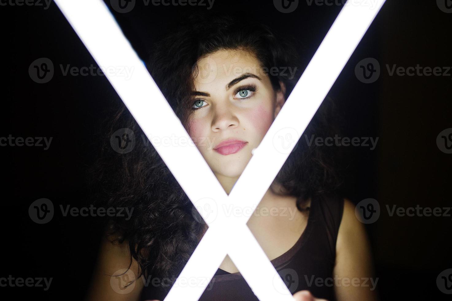Curly brunette plus size model with two long led lamp at black room. photo