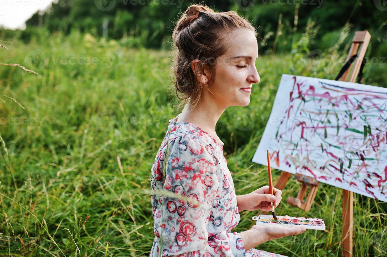 Portrait of a gorgeous happy young woman in beautiful dress sitting on the grass and painting on paper with watercolors. photo