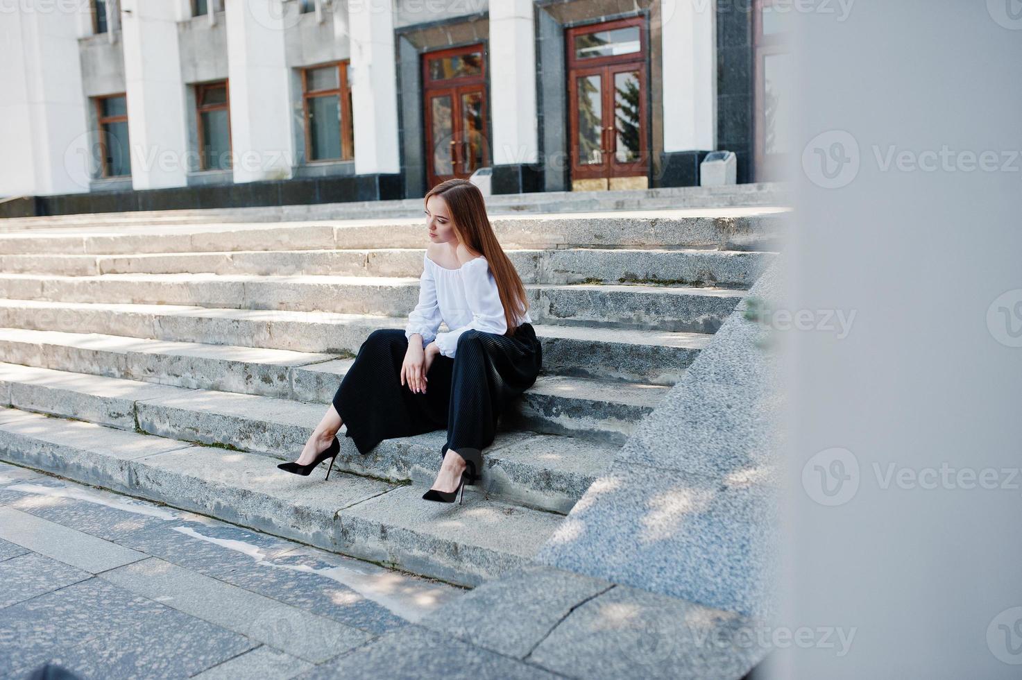 Good-looking young woman in white blouse, wide black pants and black classic high heels sitting on stairs and posing. photo