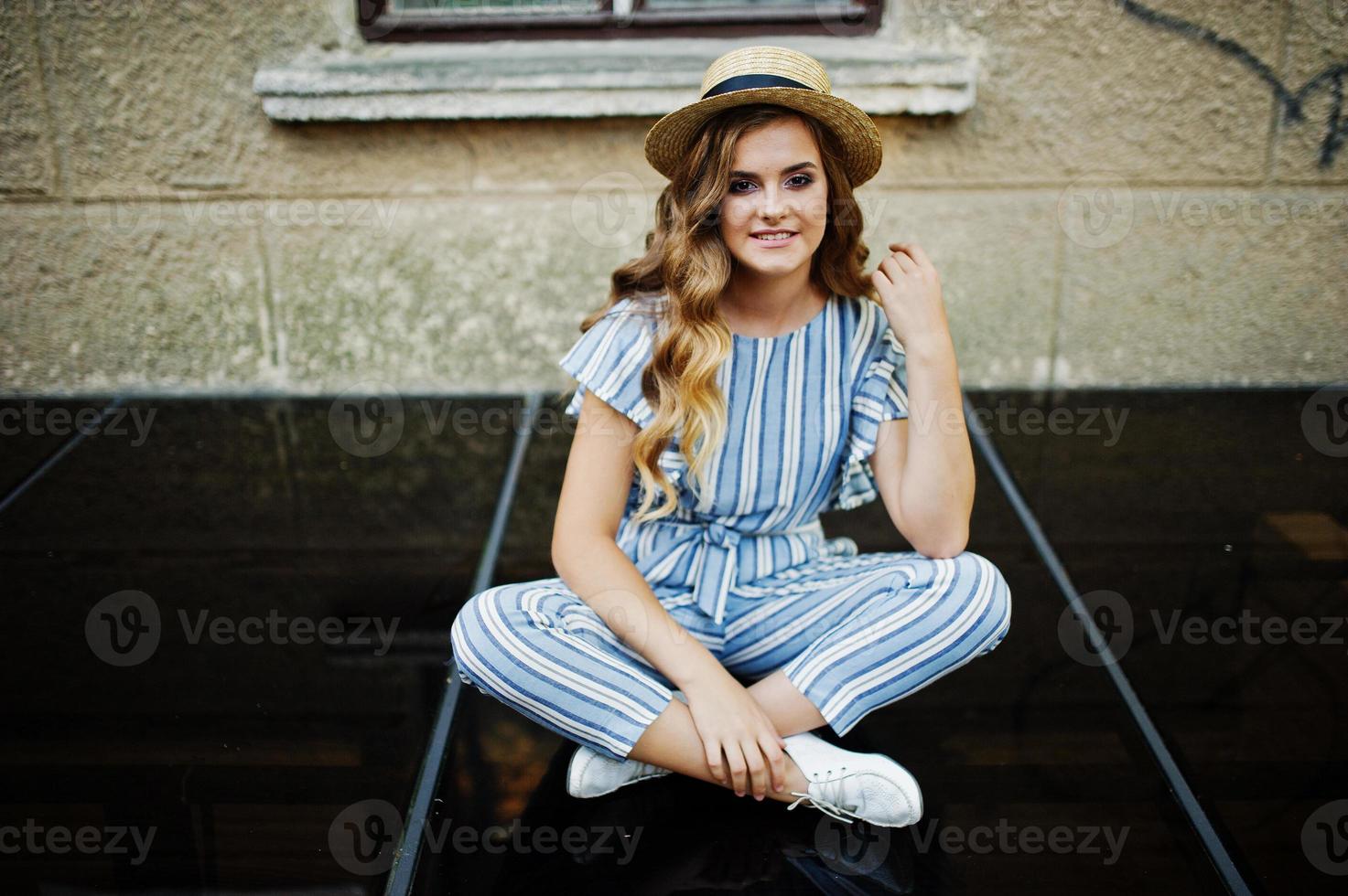 Portrait of a fabulous young woman wearing striped overall and hat sitting on black shiny surface next to the building. photo