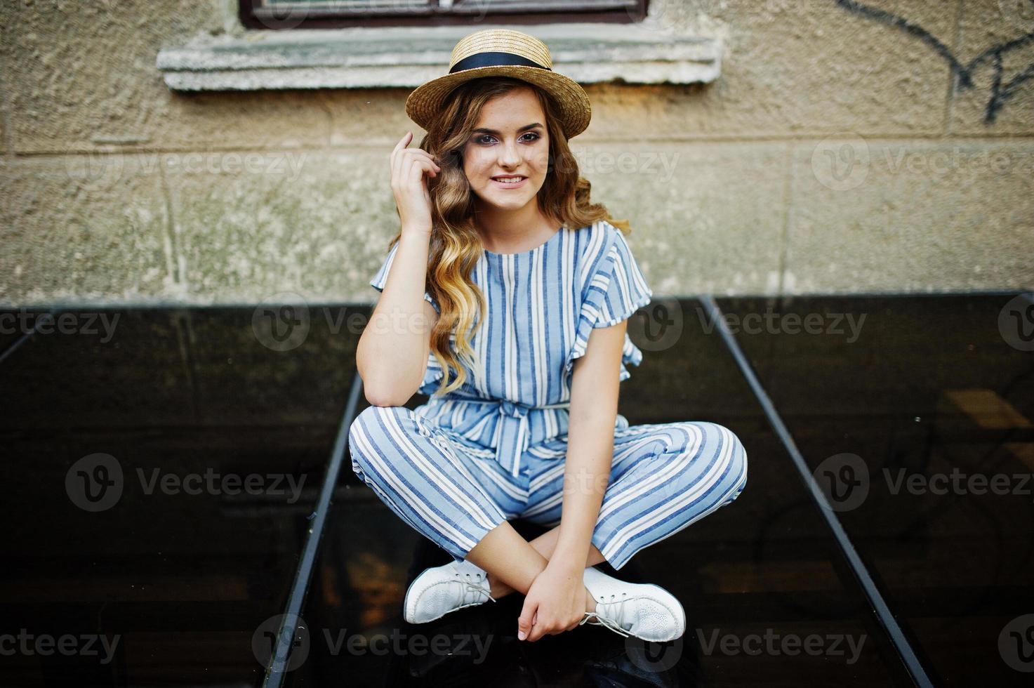 Portrait of a fabulous young woman wearing striped overall and hat sitting on black shiny surface next to the building. photo