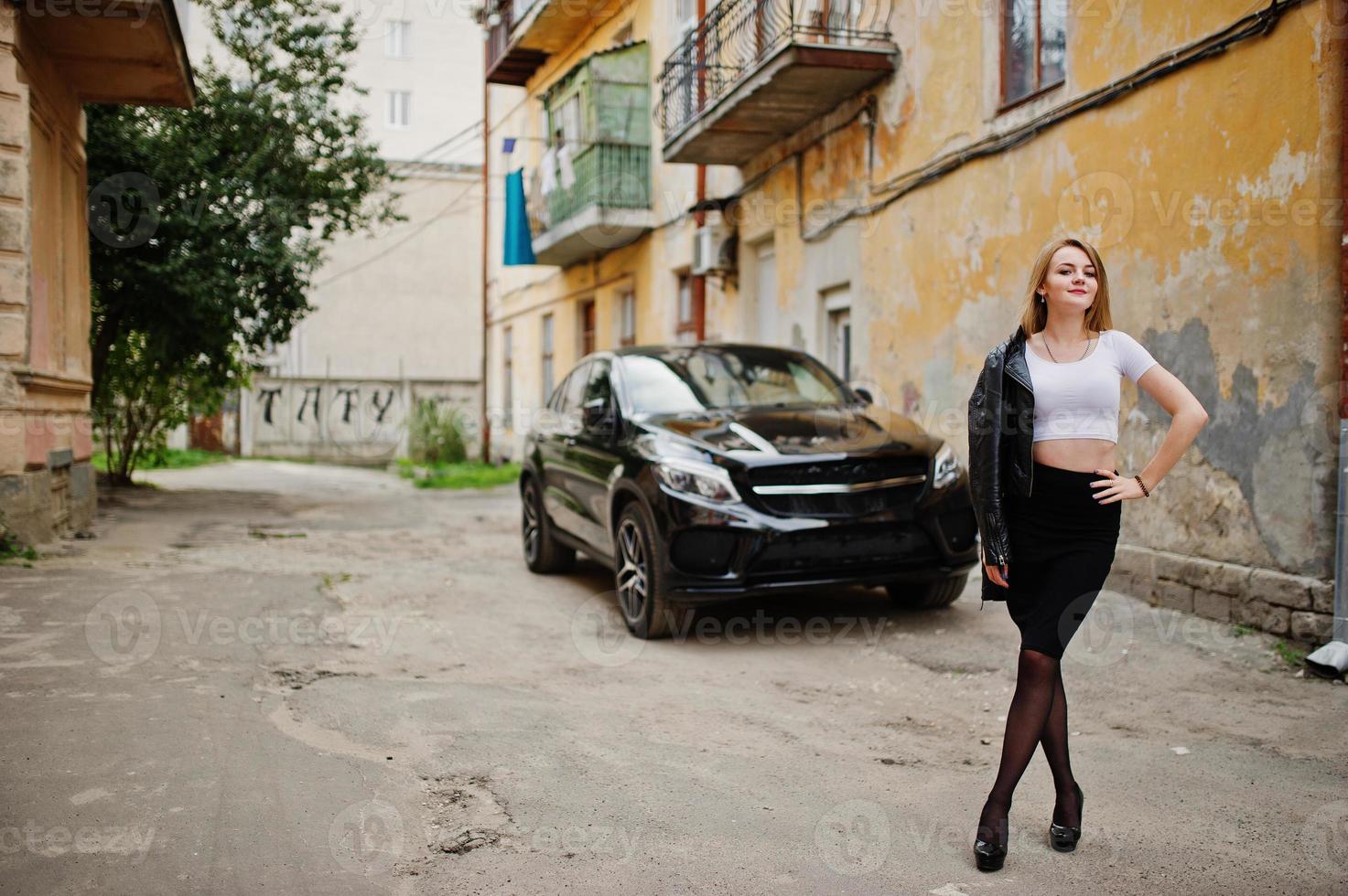 Elegant blonde girl wear on black leather jacket posing at streets of town background luxury car. photo