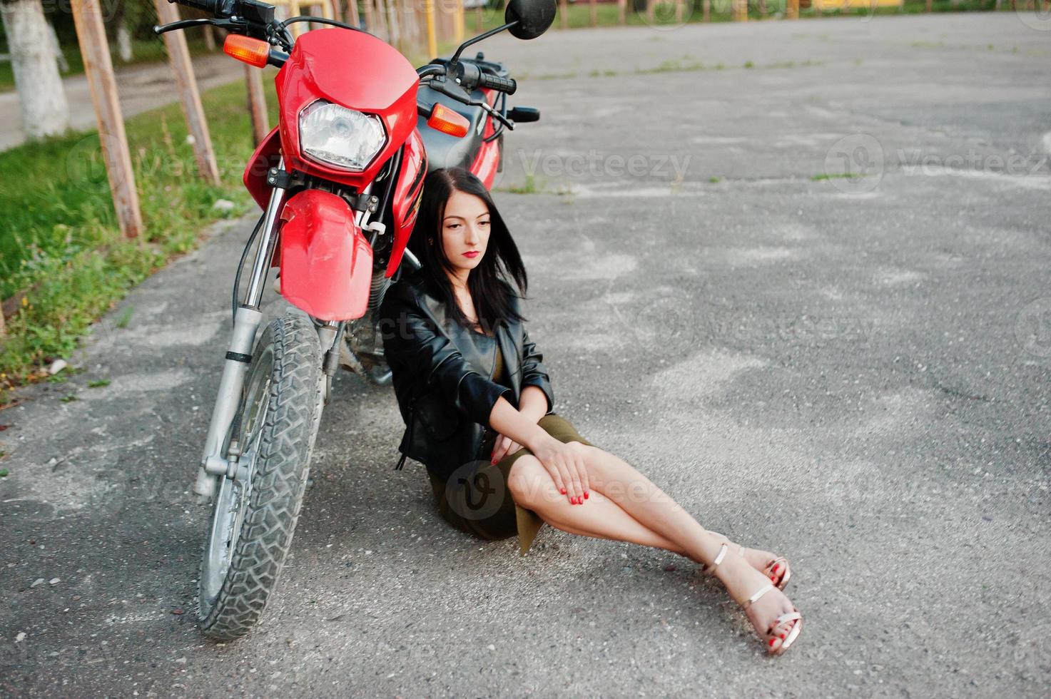 retrato de una mujer genial e impresionante con chaqueta de cuero negro sentada junto a una bicicleta roja genial. foto