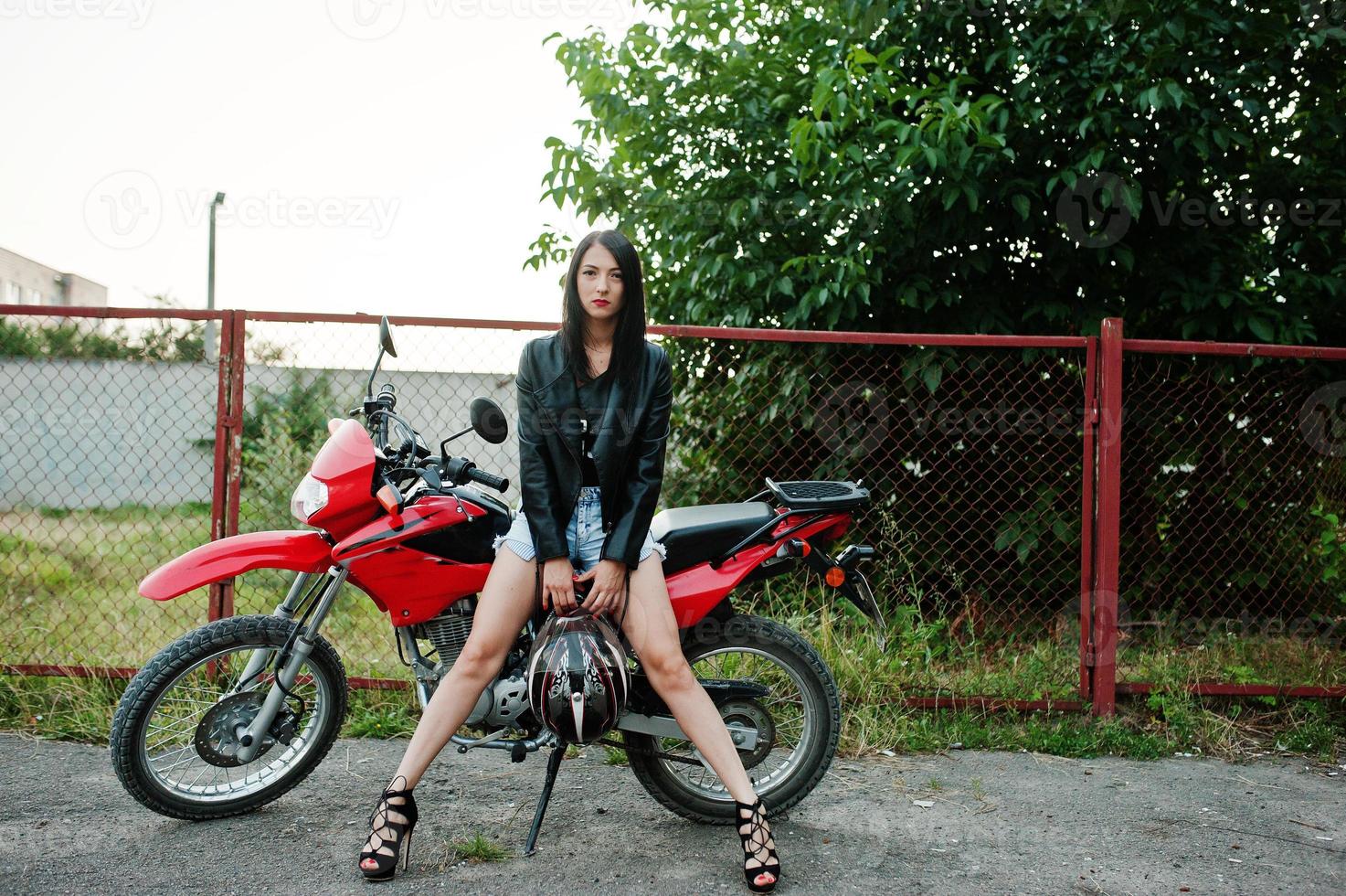 retrato de una mujer genial e impresionante con vestido y chaqueta de cuero negro sentada en una moto roja genial. foto