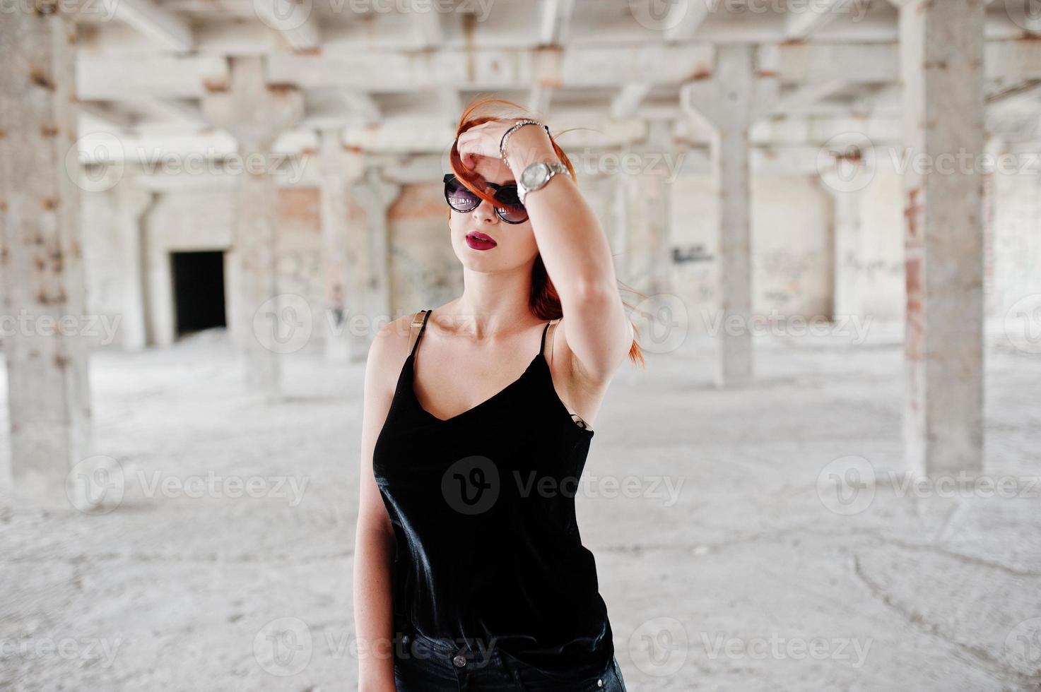chica elegante de pelo rojo con gafas de sol vestidas de negro, contra un lugar abandonado. foto