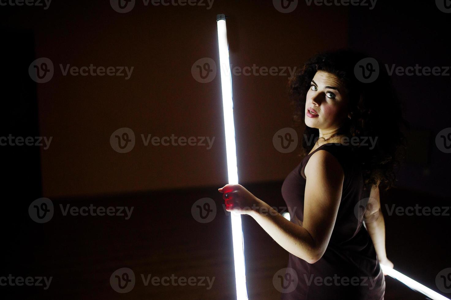 Curly brunette plus size model with two long led lamp at black room. photo
