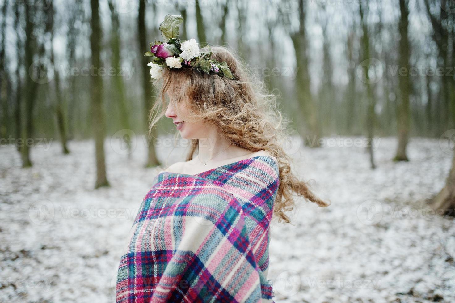 Curly cute blonde girl with wreath in checkered plaid at snowy forest in winter day. photo