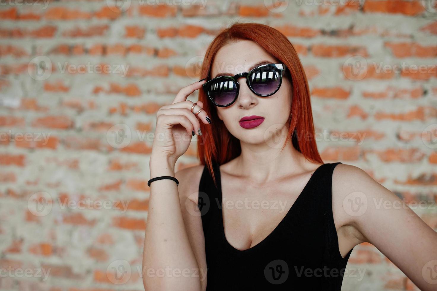 Red haired stylish girl in sunglasses wear in black, against abadoned place with brick walls. photo