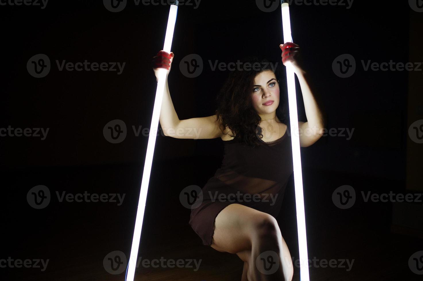 Curly brunette plus size model with two long led lamp at black room. photo