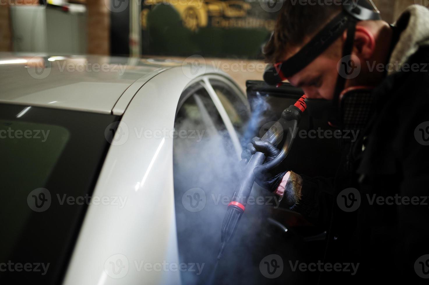 Man in uniform and respirator, worker of car wash center, cleaning car interior with hot steam cleaner. Car detailing concept. photo