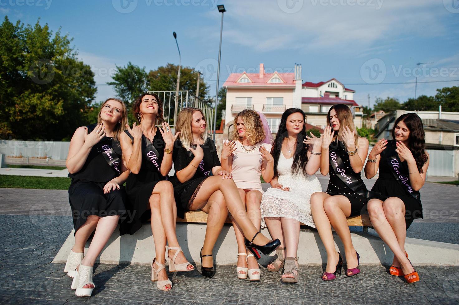 Group of 7 girls wear on black and 2 brides sitting on bench and having fun at hen party. photo