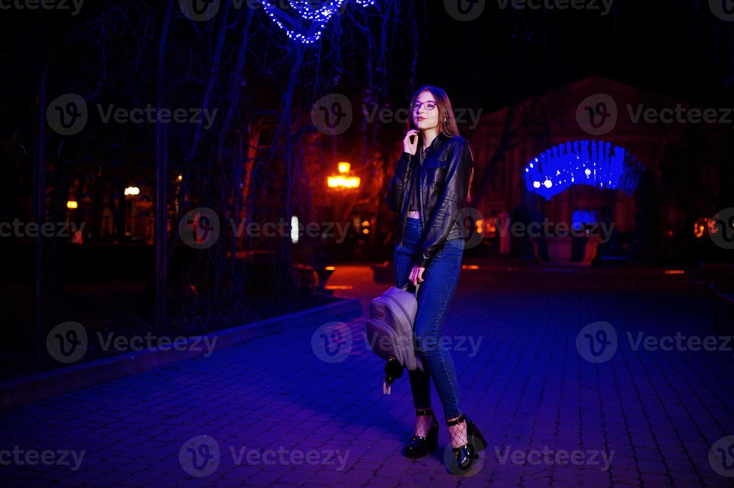 Night portrait of girl model wear on glasses, jeans and leather jacket, with backpack in hands, against blue lights garland of city street. photo