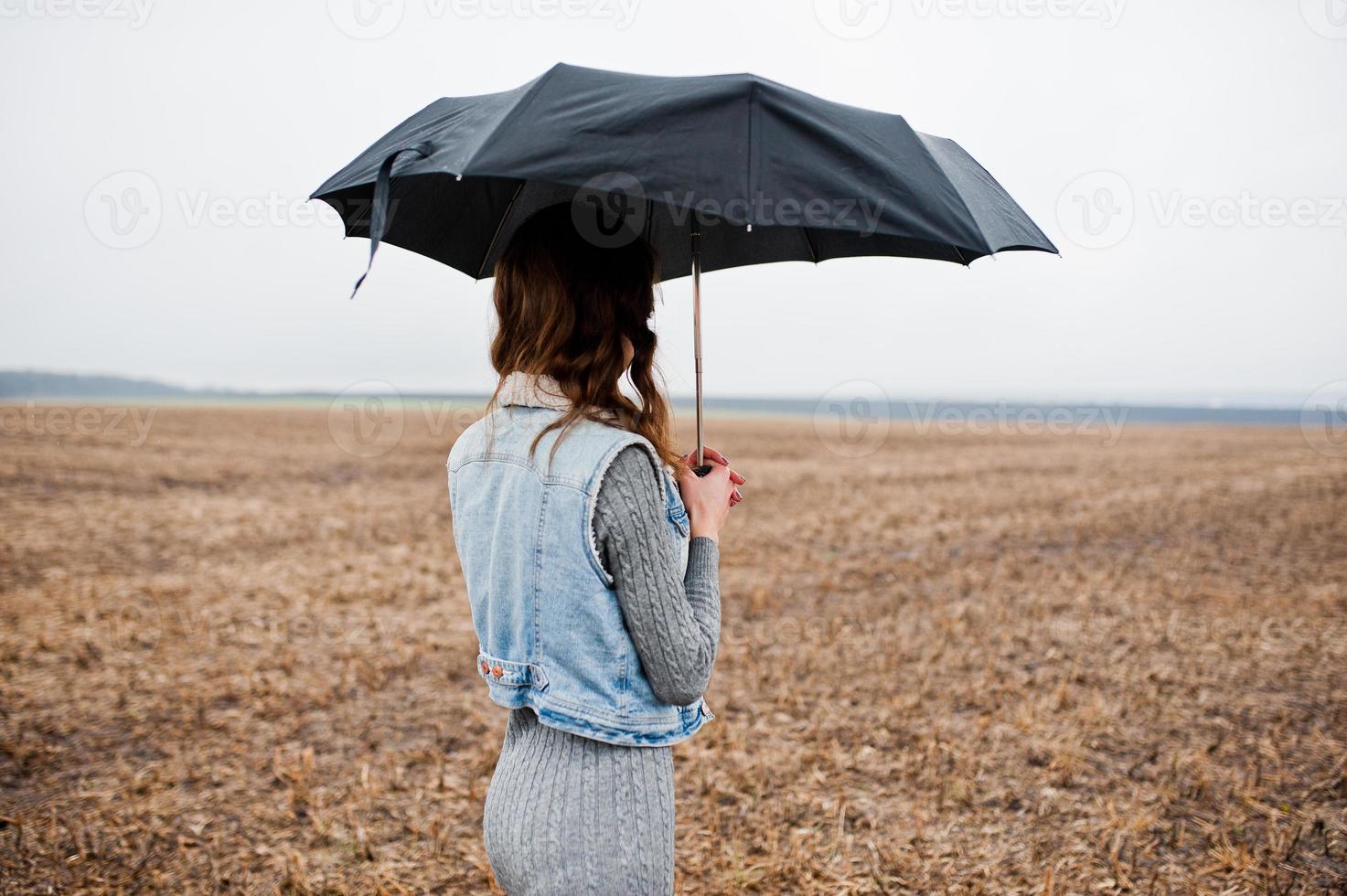 Portrait of brunette curly girl in jeans jacket with black umbrella at field. photo