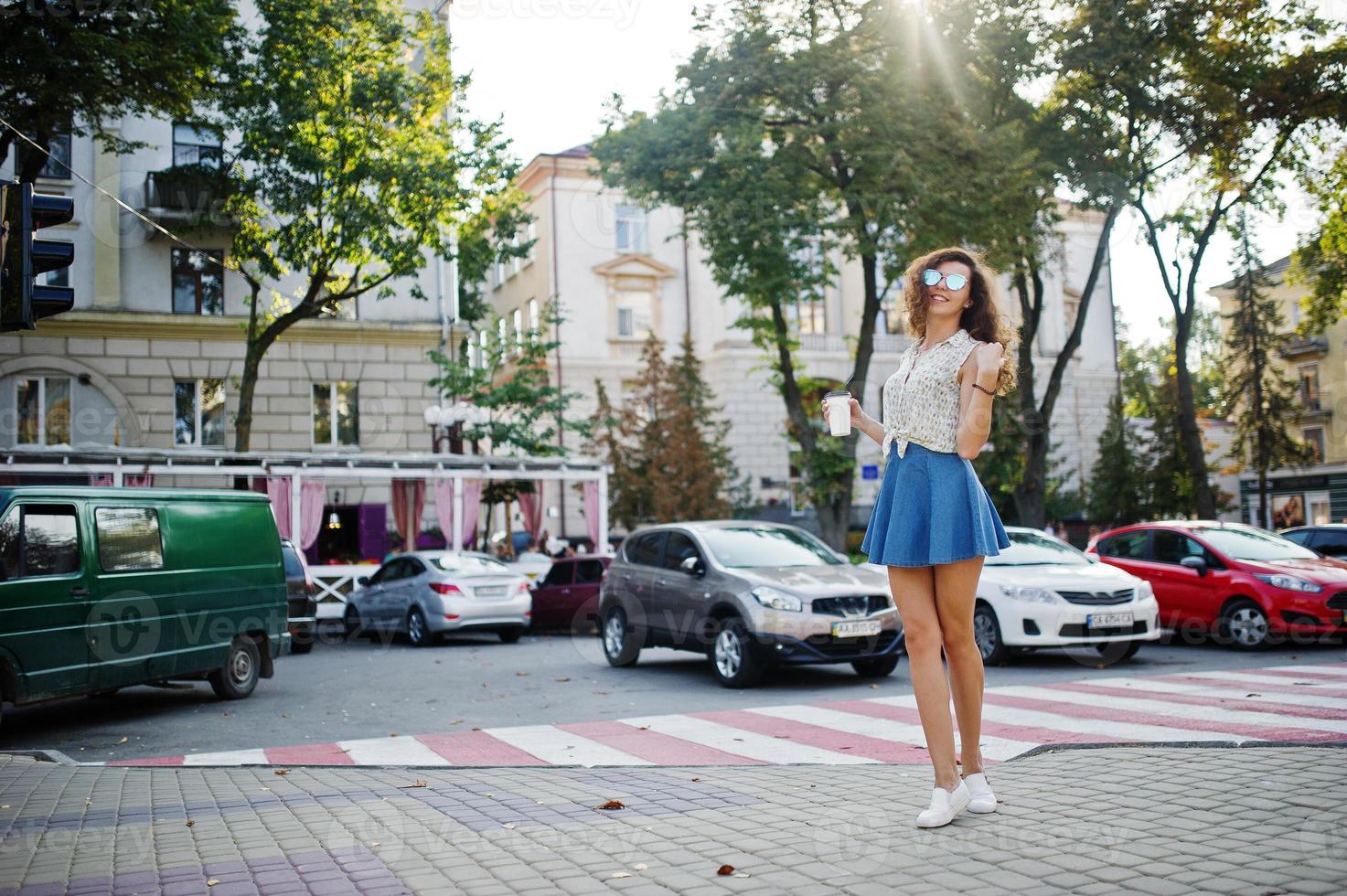 Curly stylish girl wear on blue jeans skirt, blouse and glasses posed at street of city with cup of coffee. photo