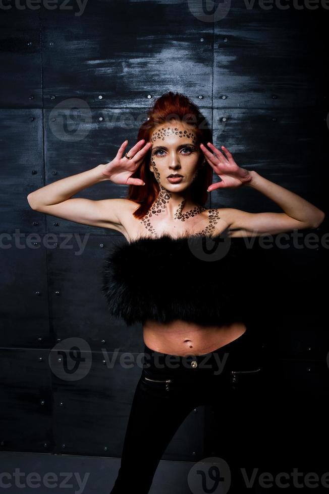 Fashion model red haired girl with originally make up like leopard predator against steel wall. Studio portrait. photo