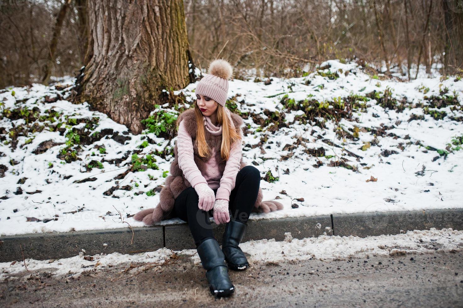 Stylish girl in fur coat and headwear at winter day on sitting border ofroad. photo