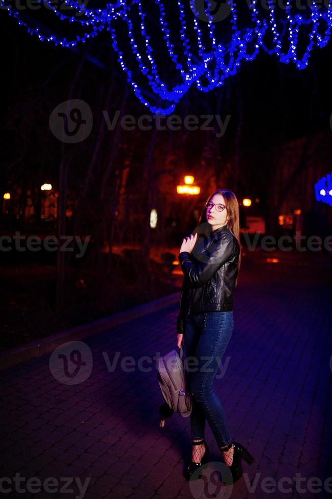 Night portrait of girl model wear on glasses, jeans and leather jacket, with backpack in hands, against blue lights garland of city street. photo