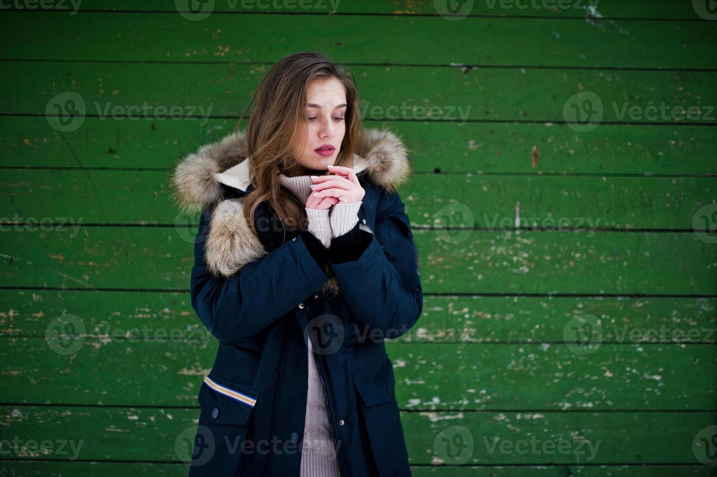 Beautiful brunette girl in winter warm clothing. Model on winter jacket against green wooden background. photo