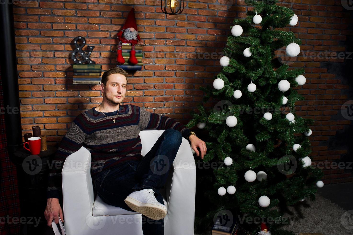 retrato de estudio de un hombre con un libro sentado en una silla contra un árbol de navidad con adornos. foto