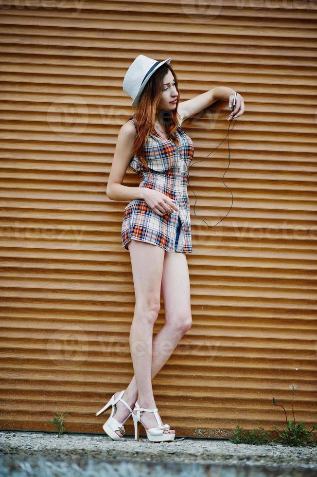 Amazing long legs with hig heels girl wear on hat against shutter listening music from headphones of mobile phone. photo