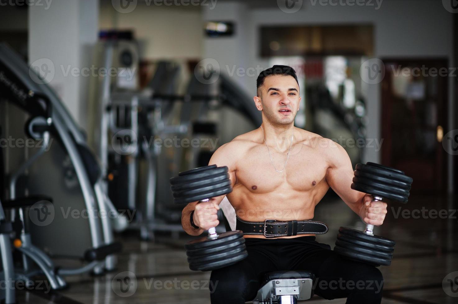 Muscular arab man training in with dumbbells modern gym. Fitness arabian men with naked torso doing workout . photo