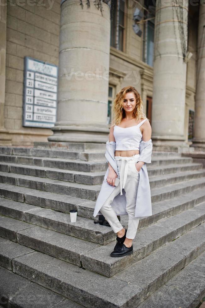 Stylish curly blonde model girl wear on white with cup of coffee posing on stairs outdoor. photo