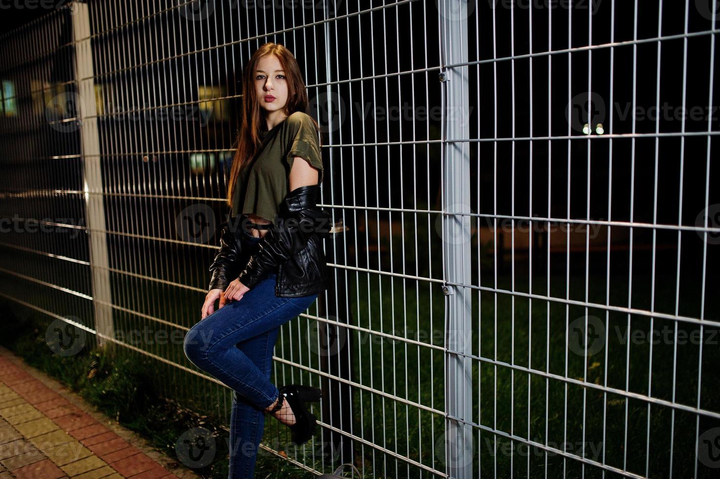 Night portrait of girl model wear on jeans and leather jacket against iron fence. photo