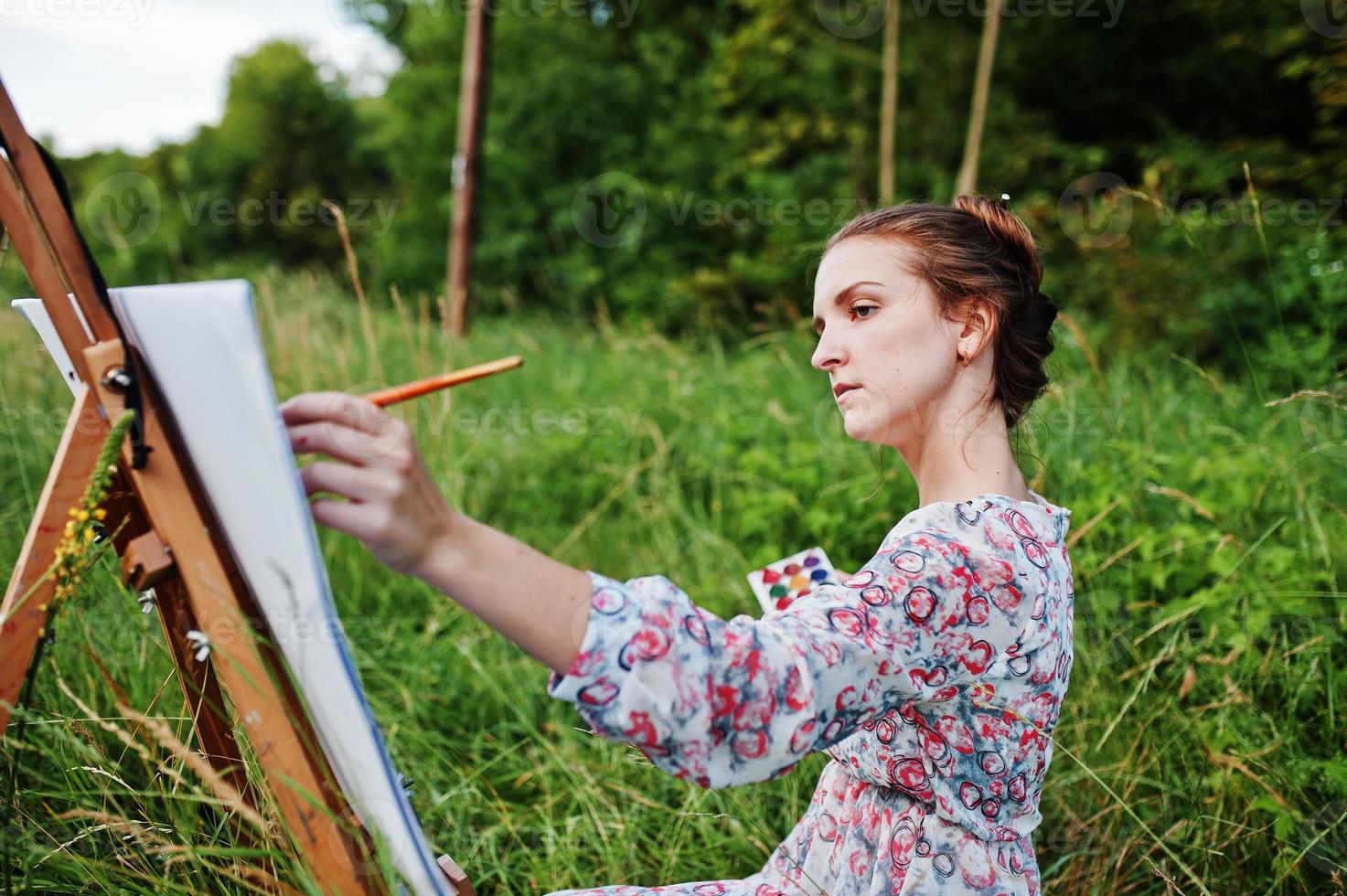 Portrait of a gorgeous happy young woman in beautiful dress sitting on the grass and painting on paper with watercolors. photo
