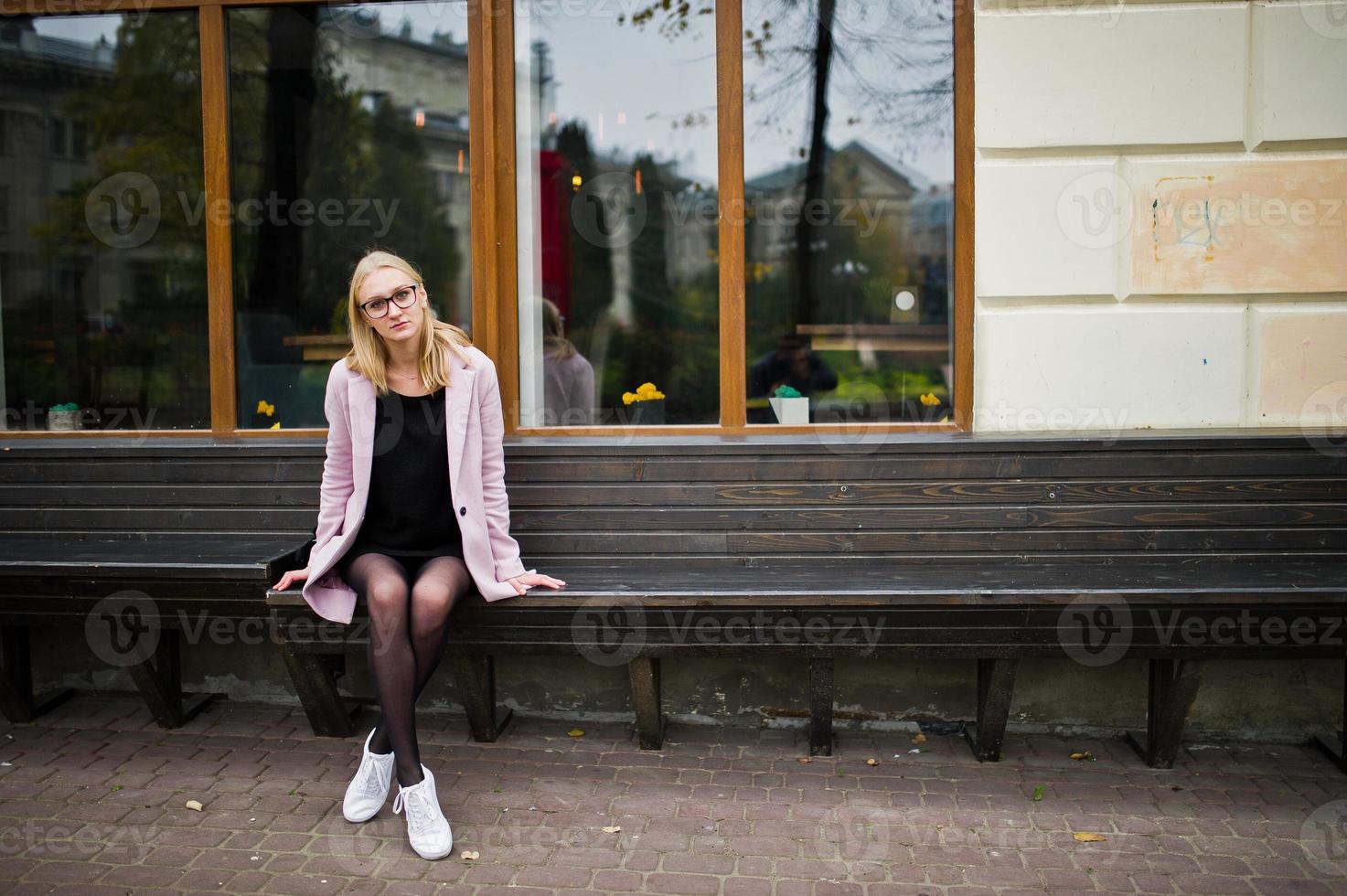 Blonde girl at glasses and pink coat, black tunic sitting on bench at street. photo